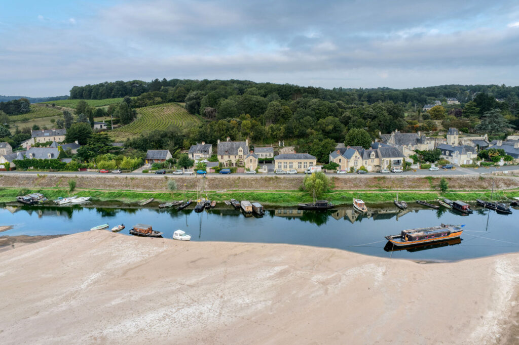 Le village du Thoureil, dans le département du Maine-et-Loire, pris depuis un drone. un banc de sable occupe le premier plan, en bas de l'image. De nombreux bateaux sont amarrés le long du quai que surplombent des maisons cossues en tuffeau et ardoise. Derrière le village, on aperçoit quelques parcelles agricoles et des massifs forestiers.